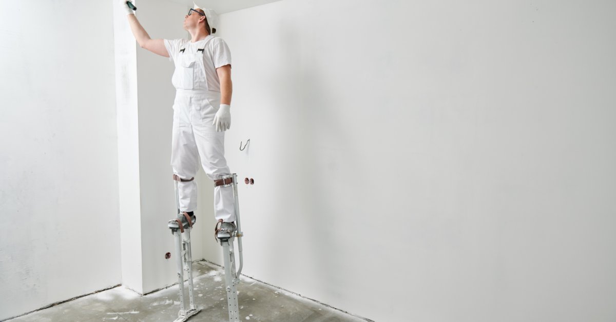 A man in white work clothes standing on drywall stilts and painting a ceiling with a roller in an unfinished room.