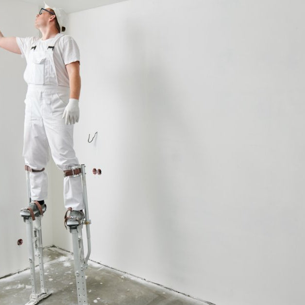 A man in white work clothes standing on drywall stilts and painting a ceiling with a roller in an unfinished room.