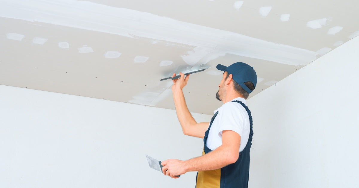 A uniformed worker applies putty to the joints of drywall sheets on a ceiling, ensuring a smooth and even finish.