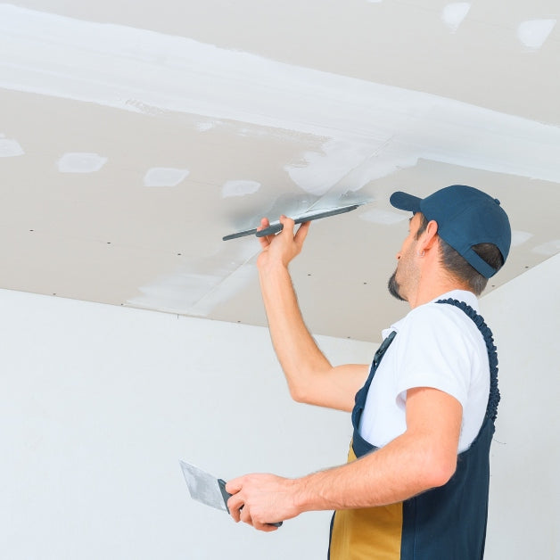 A uniformed worker applies putty to the joints of drywall sheets on a ceiling, ensuring a smooth and even finish.