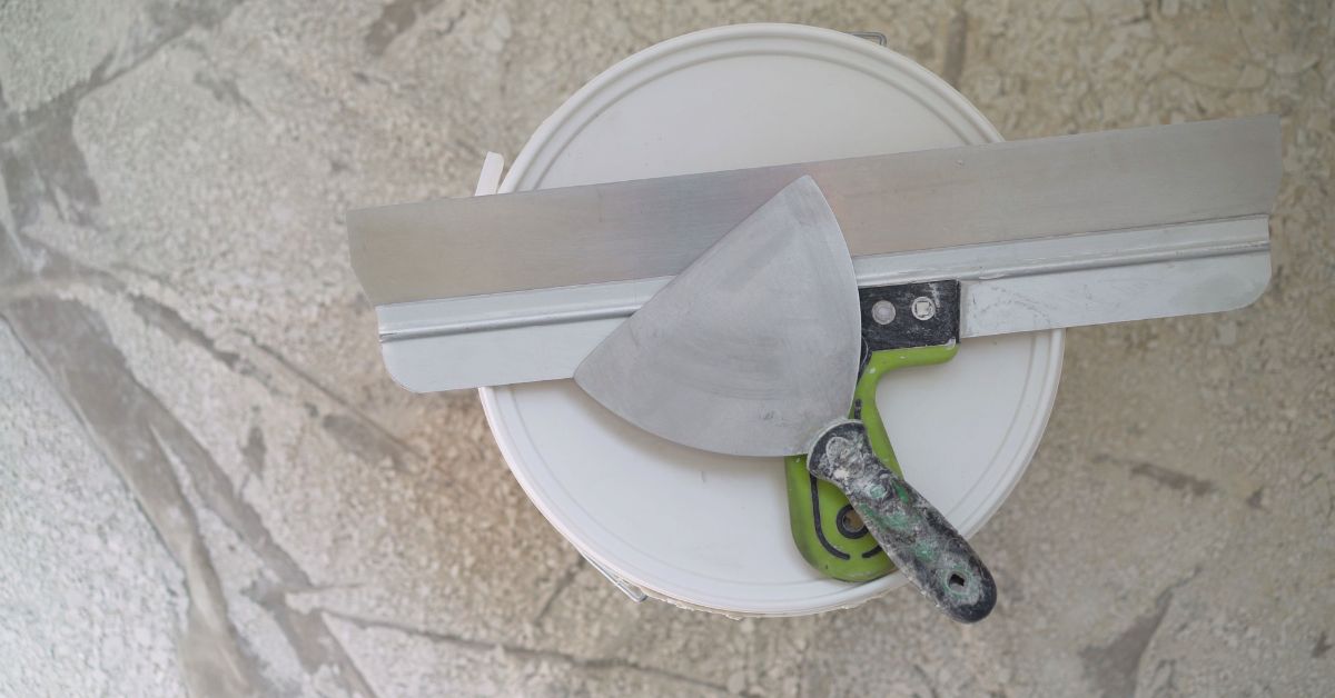 A top-down shot of two slightly used drywall knives—one large and one small—set on top of a white bucket.