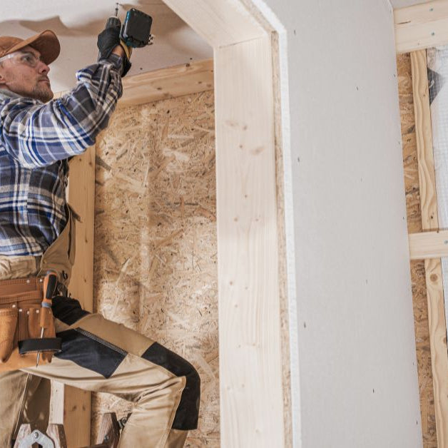 A construction contractor attaches drywall to a wooden frame using a drill driver while standing on a ladder.