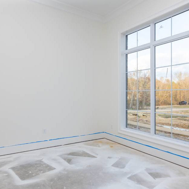 A house under construction with fresh drywall primer and painters tape on the walls. A window showcases a bright sunny day.