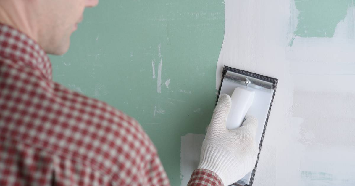 A white male wearing a red flannel shirt and gloves on carefully sanding a green wall with drywall mud.