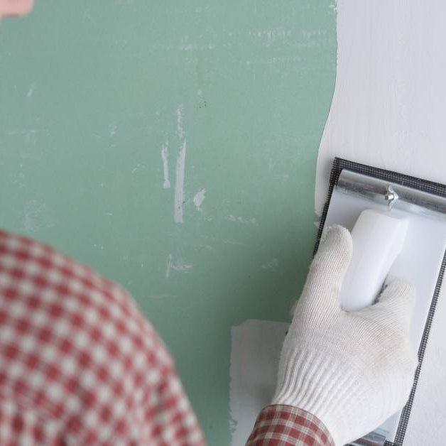 A white male wearing a red flannel shirt and gloves on carefully sanding a green wall with drywall mud.