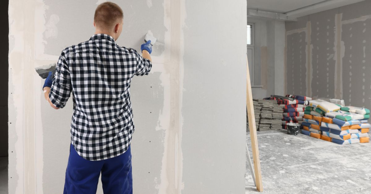 A man in a black and white gingham shirt and blue pants holding a taping knife loaded with drywall mud.