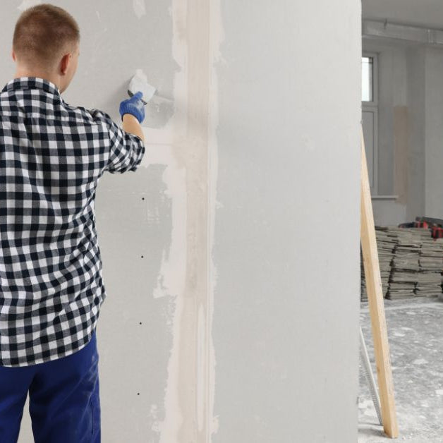 A man in a black and white gingham shirt and blue pants holding a taping knife loaded with drywall mud.