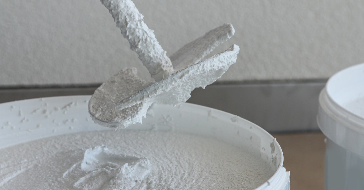 A focus shot of a drywall mixer being lifted out of a white bucket of plaster. The mixer is coated in white plaster mix.