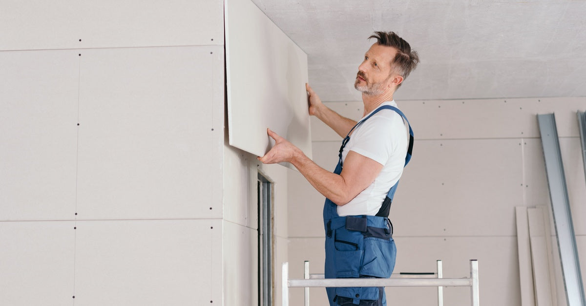 A man wearing work overalls and a white T-shirt is standing on a drywall bench and putting up sheets of drywall.