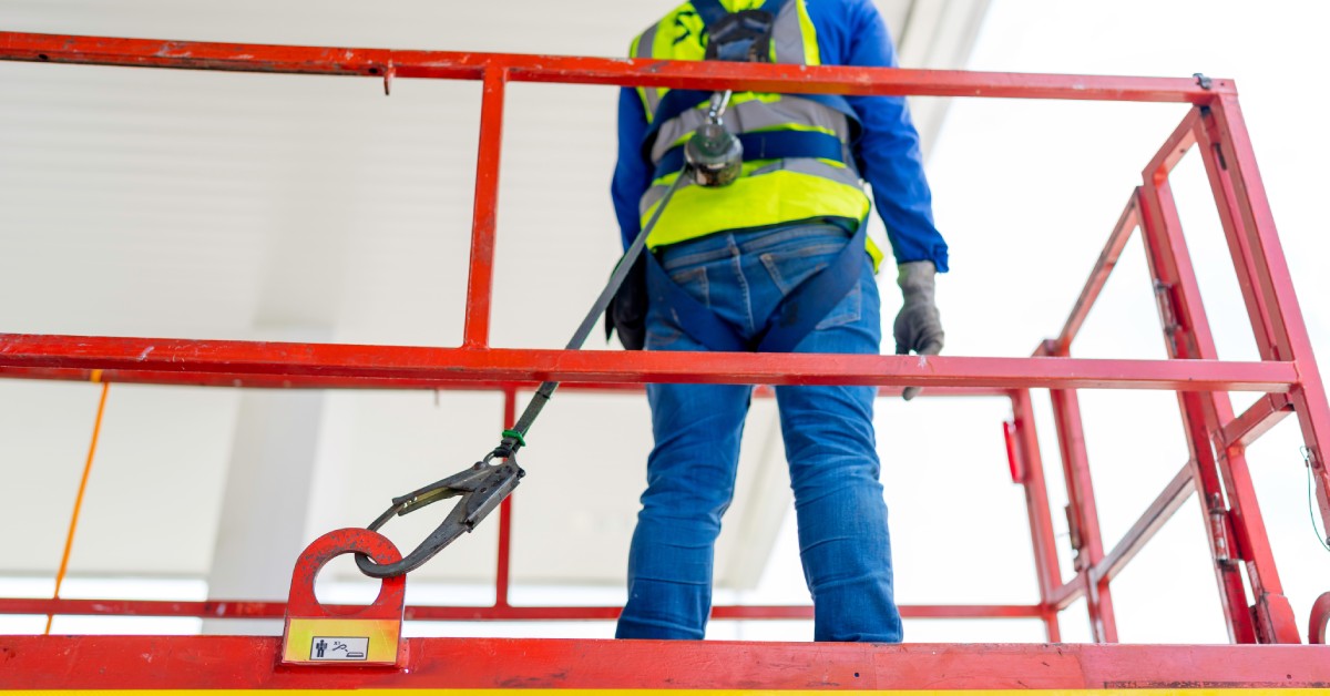 A man wearing construction gear, secured by a rope around his waist, standing on a lift raised near the ceiling.