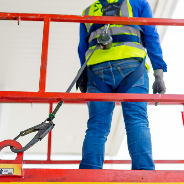 A man wearing construction gear, secured by a rope around his waist, standing on a lift raised near the ceiling.