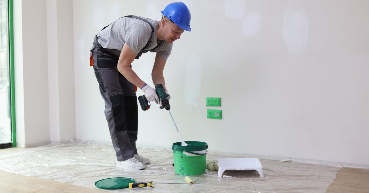A person in a hard hat and overalls using a power drill with a paddle attachment to mix materials in a green bucket.
