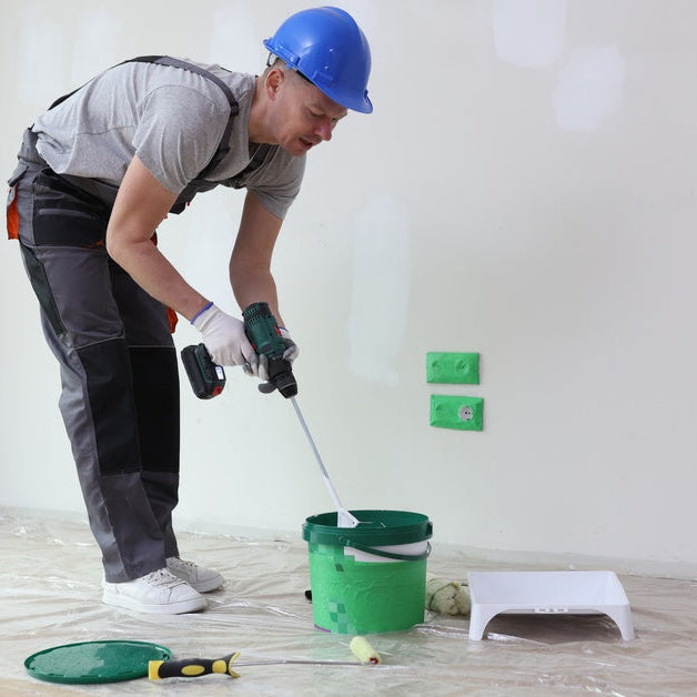 A person in a hard hat and overalls using a power drill with a paddle attachment to mix materials in a green bucket.