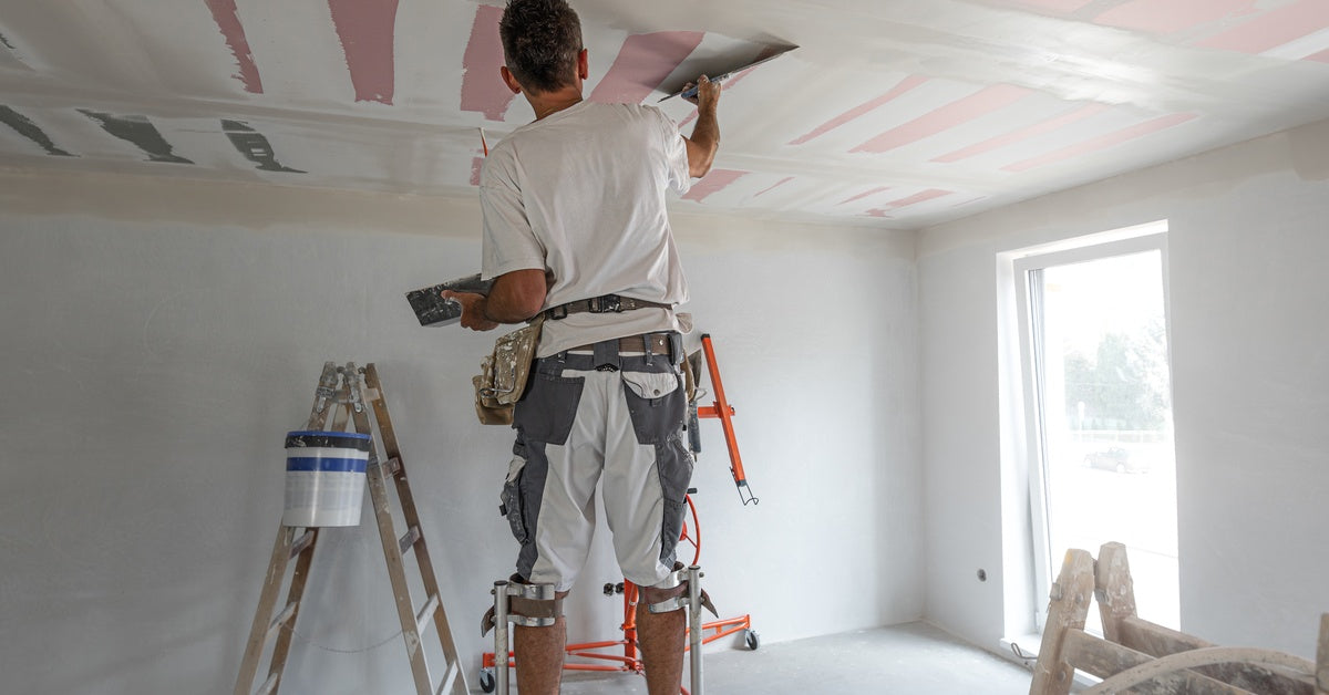 A worker on drywall stilts applying joint compound to a ceiling with a wide taping knife in a new build.