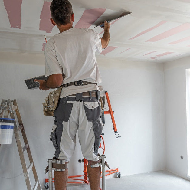 A worker on drywall stilts applying joint compound to a ceiling with a wide taping knife in a new build.