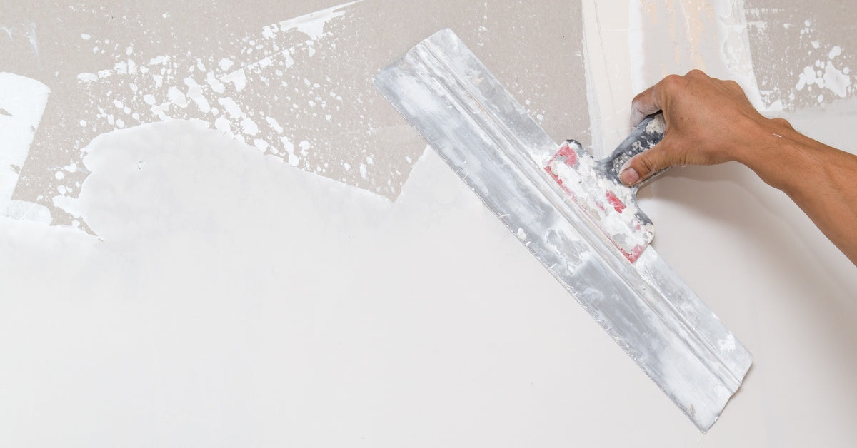 A gloved hand using a trowel to spread gray plaster across a wall during indoor surface repair work.