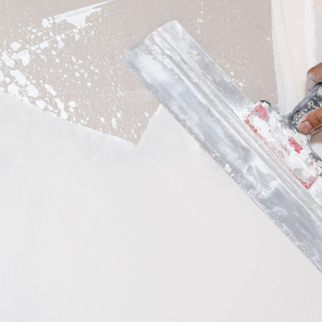 A gloved hand using a trowel to spread gray plaster across a wall during indoor surface repair work.