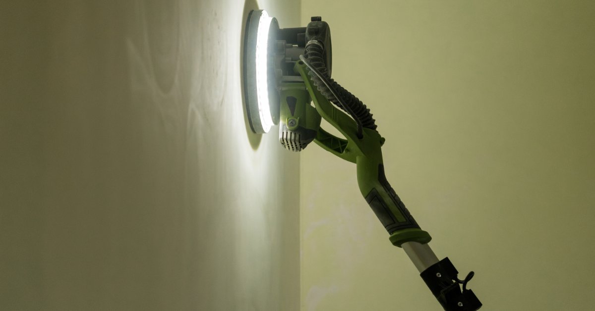 A green pole sander smoothing a white interior wall while a person holds the tool near a bright corner.
