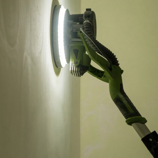 A green pole sander smoothing a white interior wall while a person holds the tool near a bright corner.