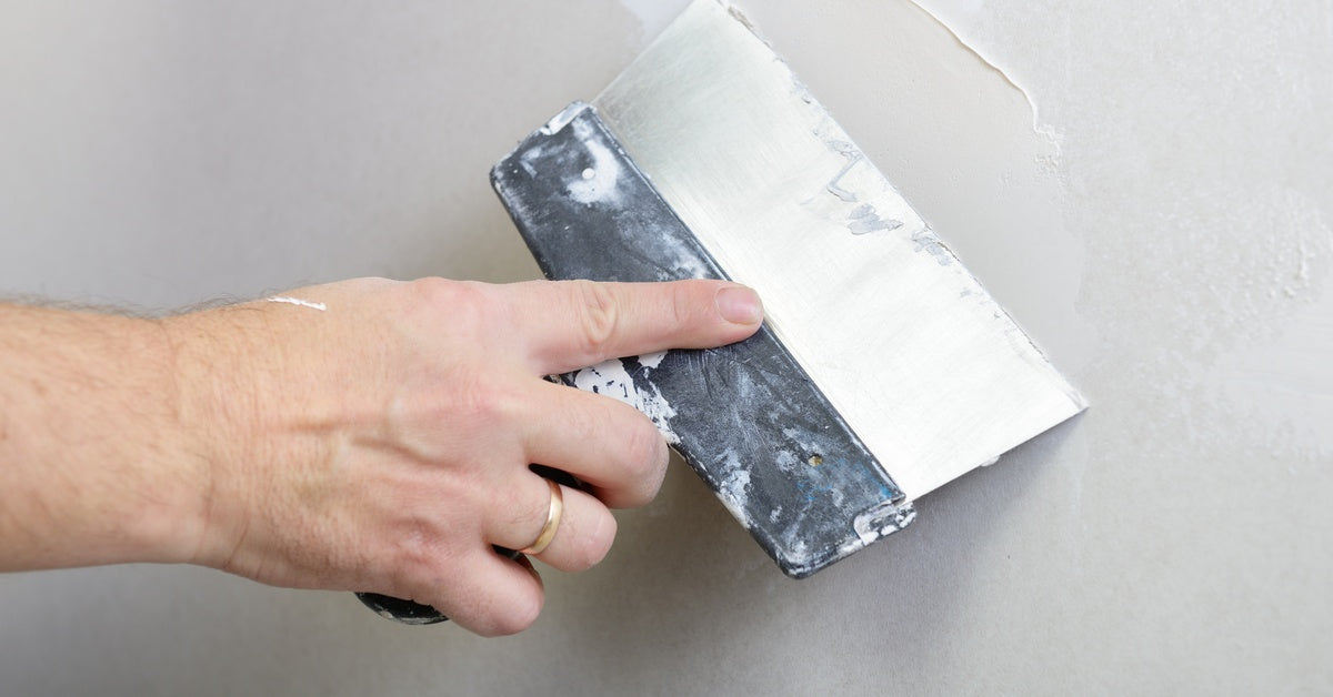 A close-up of a hand holding a wide putty knife and smoothing compound evenly across a gray wall surface.