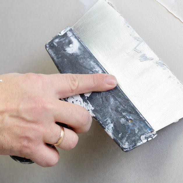 A close-up of a hand holding a wide putty knife and smoothing compound evenly across a gray wall surface.