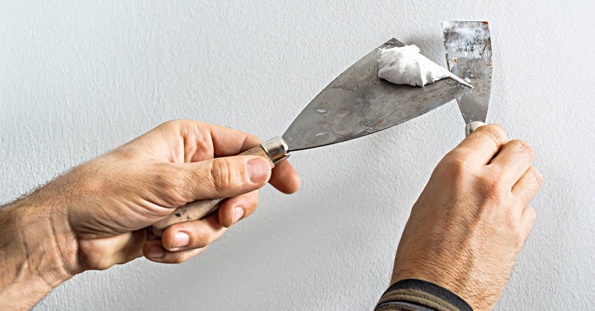 Hands holding a putty knife and scraper with white filler against a textured wall during repair work.