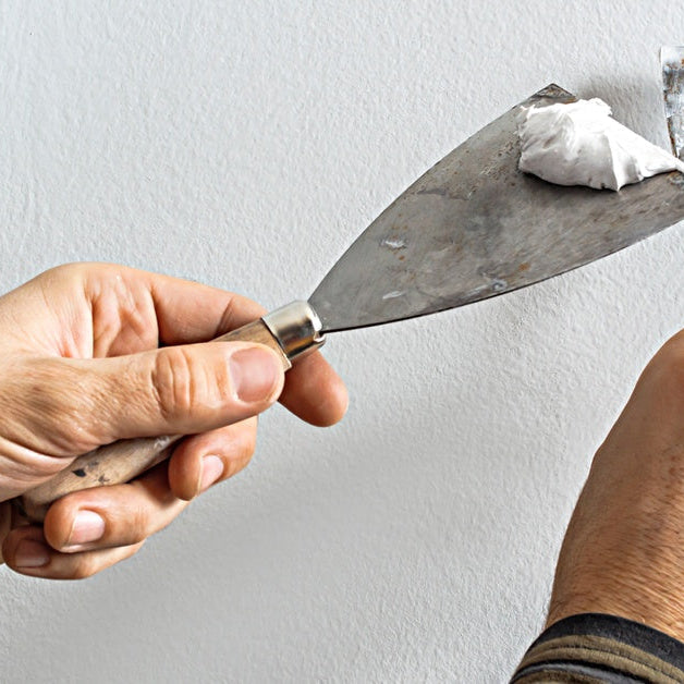 Hands holding a putty knife and scraper with white filler against a textured wall during repair work.